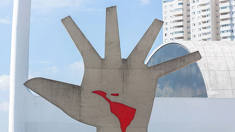 Foto de una figura metálica que representa una mano gigante de varios metros. En medio de la mano se ve en rojo el contorno de América Latina. 