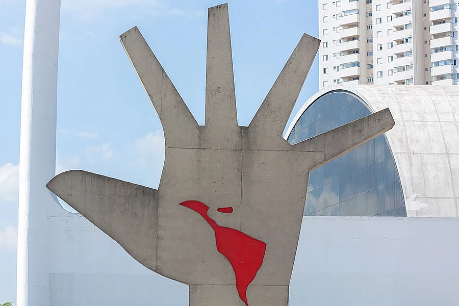 Foto de una figura metálica que representa una mano gigante de varios metros. En medio de la mano se ve en rojo el contorno de América Latina. 