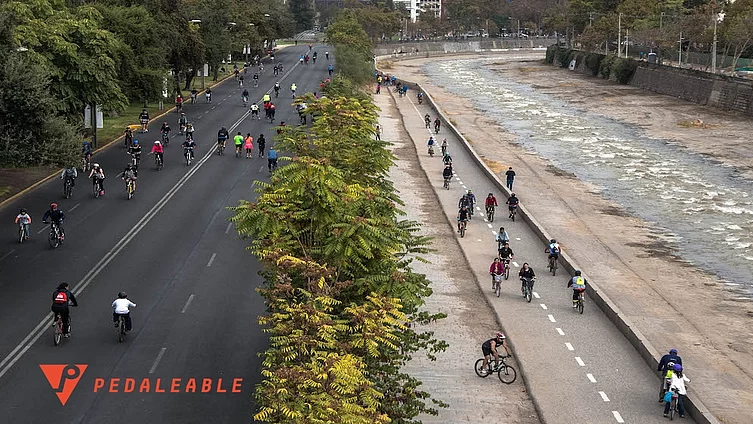 Fotografía del río Mapocho en Santiago en un día de 'Mapocho pedaleable'. Al lado izquierdo se ve una calle ancha sin autos pero con muchos ciclistas. Al medio se ven arbustos verdes. Más a la derecha se ve una ciclovía al lado del río también con ciclistas y a la derecha el lecho del río Mapocho.