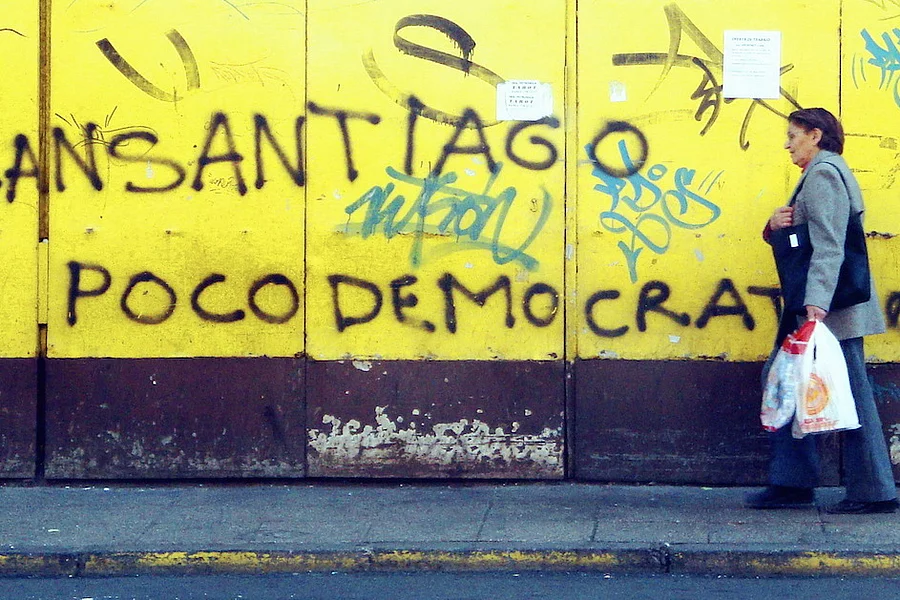 Grafiti en un muro amarillo que dice 'Transantiago poco democrático'. Desde el lado derecho pasa una mujer adulta mayor con un bolso pesado.