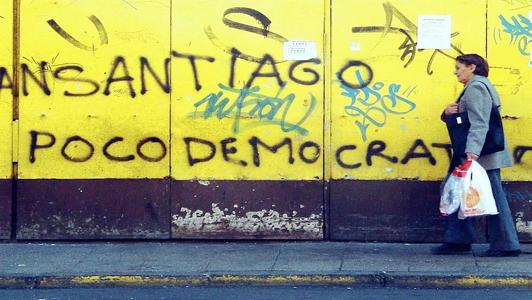 Grafiti en un muro amarillo que dice 'Transantiago poco democrático'. Desde el lado derecho pasa una mujer adulta mayor con un bolso pesado.