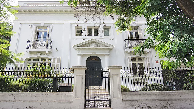Foto de la casa de la FES Chile. Una casona antigua blanca detrás de unas rejas y arbustos intermedio.